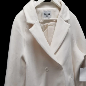 Vit kappa från NA-KD - Säljer en superfin vit kappa från NA-KD i storlek 38 längden från axel måttat 85 cm. Den har en klassisk design med dubbelknäppning och långa ärmar. Perfekt för både höst och vår! Kappan är i nyskick och har en stilren look som passar till alla outfits. 🕊️