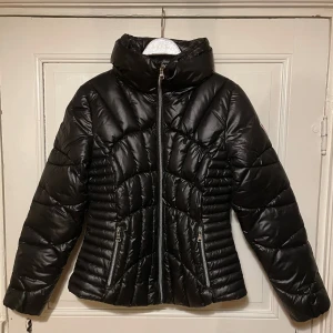 GUESS pufferjacka svart - Säljer en elegant svart pufferjacka från GUESS i storlek Medium. Jackan är helt ny och aldrig använd, perfekt för kalla dagar med sin quiltade design och höga krage som håller dig varm och bekväm.  