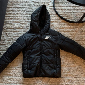 Svart pufferjacka från Nike - Säljer en svart pufferjacka från Nike. Den är perfekt för kalla dagar med sin vadderade design och huva. Jackan har en dragkedja framtill och är långärmad. Nike-loggan syns tydligt på bröstet. 