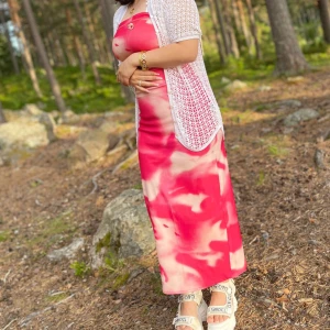 Rosa och vit långklänning - Säljer en superfin rosa långklänning med ett unikt mönster. Klänningen är ärmlös och perfekt för sommardagar. Den har en bekväm passform och är gjord i ett mjukt material. Perfekt för en avslappnad dag i naturen eller en sommarfest!