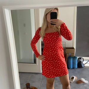 Röd blommig klänning - Säljer en superfin röd klänning med blommigt mönster. Den har en tight passform och är långärmad, perfekt för både vår och sommar. Klänningen är kort och har en fyrkantig urringning som ger en söt touch. Perfekt för både vardag och fest!