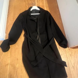 Svart kappa  - Snygg svart kappa  i bra skick. Den har ett bälte i midjan för en elegant look och är perfekt för höst och vår. Långärmad och i en klassisk stil som passar till det mesta. Perfekt för både vardag och fest!