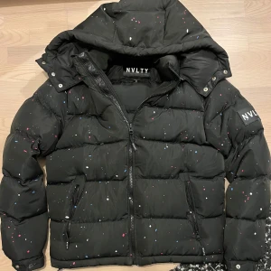 Svart pufferjacka från NVLTY - Säljer en svart pufferjacka från NVLTY i storlek M. Jackan har ett coolt färgstänk-mönster och är perfekt för kalla dagar. Den har en dragkedja framtill och praktiska fickor. Huvan är justerbar och jackan är i mycket bra skick. Perfekt för vintern! 
