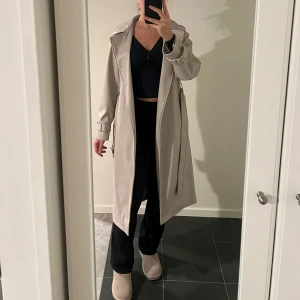 Beige/grå trenchcoat skinn med bälte - Säljer en snygg beige/grå skinn trenchcoat i mycket bra skick som är köpt från Stradivarius . Den har ett bälte i midjan och långa ärmar med justerbara spännen. Perfekt för höst och vår! Passar till både vardag och fest. Storlek dam xs.