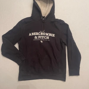 Svart fleecefodrad hoodie från Abercrombie & Fitch - Säljer en svart hoodie med riktigt skön fleece från Abercrombie & Fitch i storlek M. Den har en mysig fleecefodrad huva och tryck på framsidan. Perfekt för kalla dagar och avslappnade stunder. Den är storlek M men passar även S