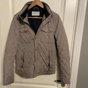 Beige quiltad jacka från Zara Man - Säljer en snygg beige quiltad jacka från Zara Man. Perfekt för höst och vår med sin stilrena design och praktiska fickor. Jackan har dragkedja och knappar framtill samt långa ärmar, inbyggd luva i nacken. Den är i mycket bra skick och passar perfekt för s