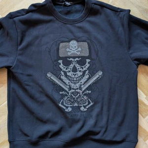 Svart tröja från Philipp Plein - Säljer en svart tröja från Philipp Plein med ett coolt dödskallemotiv och korsade benknotor på framsidan. Tröjan har en liten passform, mer som en XL eller oversize L och är perfekt för höst och vinter. Den är i mycket bra skick och har en unik design med små detaljer i mönstret med kristall liknande ploppar.