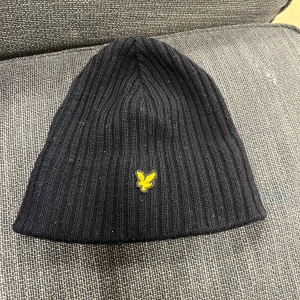 Svart ribbad mössa från Lyle & Scott - Säljer en svart ribbad mössa från Lyle & Scott med deras ikoniska gula logga framtill. Mössan är perfekt för kyliga dagar och har en stilren design som passar till det mesta. Den är i mycket bra skick och redo för nya äventyr!