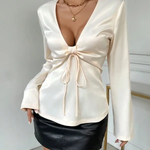 Krämvit satinblus med knytning - Elegant krämvit blus i satin med djup v-ringning och knytning framtill. Långärmad och perfekt för både fest och vardag. Den mjuka satinfinishen ger en lyxig känsla.