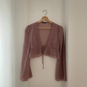 Beige blus med knytband - Säljer en superfin lila blus med långa ärmar och knytband framtill. Den har en v-ringad design och volangdetaljer vid nederkanten. Perfekt för både vardag och fest! 💫