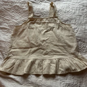 Beige linnetopp från Name It - Säljer en söt beige linnetopp från Name It i storlek 146-152. Toppen har breda axelband och en volangdetalj längst ner som ger en fin touch. Perfekt för sommardagar! 🌞