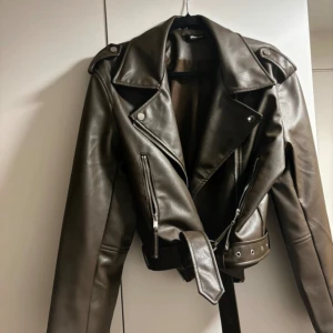 Grön-brun skinnjacka med bälte - Säljer en snygg brun skinnjacka i mycket bra skick. Den har en cool bikerstil med dragkedjor och ett bälte i midjan. Perfekt för höst och vår! Jackan är långärmad och har en klassisk krage. Passar perfekt för en edgy look.