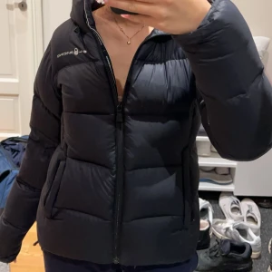 Svart dunjacka från Sail Racing - Säljer en svart dunjacka från Racing i mycket bra skick. Jackan har en dragkedja framtill och är långärmad. Den är både varm och stilren, med en normal passform. Den har små hål på ärmarna som knappt syns, kom privat för bilder, kan sänka priset vid snabb affär. Kan byta mot annan jacka