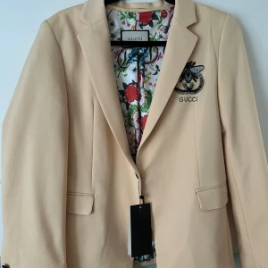 Beige kavaj från Gucci - Säljer en snygg beige kavaj från Gucci i nytt skick. Den har ett coolt broderat bi-motiv på ryggen och ett färgglatt blommigt foder. Kavajen har en klassisk passform med två fickor fram och en knapp för stängning. Perfekt för både fest och vardag!