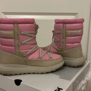 Rosa och beige vinterstövlar från Helly Hansen - Säljer ett par supermysiga rosa och beige vinterstövlar från Helly Hansen. De har en vadderad design med snörning framtill och är perfekta för kalla vinterdagar. Stövlarna är i mycket bra skick och redo för nya äventyr i snön! ❄️ orginalpris 1700kr