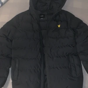 Svart pufferjacka från Lyle & Scott - Säljer en svart pufferjacka från Lyle & Scott i storlek 12/13 år. Jackan har en mysig huva och är perfekt för kalla dagar. Den är i mycket bra skick och har en dragkedja framtill. Perfekt för vintern! 🖤