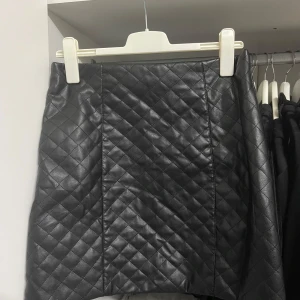 Svart quiltad kjol från Amisu - Snygg svart quiltad kjol från Amisu i storlek EU 40. Kjolen har en cool dragkedja baktill och är perfekt för höst och vinter. Den är gjord i ett skinnliknande material som ger en edgy look. Passar både till vardags och fest!