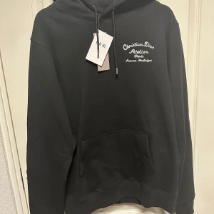Svart hoodie  - Säljer en snygg svart hoodie från med broderad text både fram och bak. Den har en klassisk passform med en stor ficka fram och justerbar huva. Perfekt för både vardag och lite finare tillfällen. Helt ny med tag kvar!
