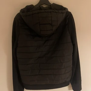 Svart jacka från J.Lindberg - Säljer en svart cardigan jacka från j.Lindberg i bra skick. Den har en dragkedja framtill och en mysig huva. Perfekt nu när det är lite kyligt ute. Jackan är stilren och passar till det mesta. Har ett litet hål i ena fickan, skriv ifall du vill ha bild! Hör även av dig  ifall du har någon fråga eller fundering!!