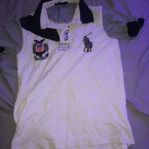 Vit och grå pikétröja från Polo Ralph Lauren - Säljer en snygg vit och grå pikétröja från Polo Ralph Lauren. Den har korta ärmar och en klassisk krage i mörkblått. Tröjan är dekorerad med broderade detaljer på framsidan. Perfekt för en avslappnad men stilren look!