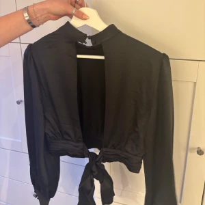 Svart långärmad topp med knyt - Snygg svart topp med långa ärmar och knytning i midjan. Perfekt för en kväll ute eller en fest. Den har en hög hals och är i ett mjukt material som ger en elegant look.