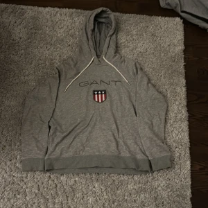 Grå hoodie från GANT - Säljer en snygg grå hoodie från GANT i storlek L i barnstorlek. Den har en klassisk design med GANT-loggan och ett coolt emblem på bröstet. Perfekt för en avslappnad stil. Huvan har vita dragsnören med GANT-detaljer. Superbekväm och stilren!