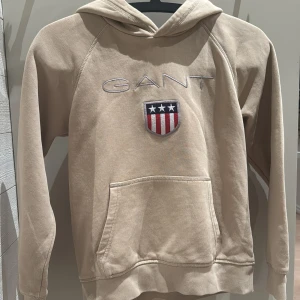 Beige hoodie från GANT - Säljer en snygg beige hoodie från GANT med broderad logga och ett coolt emblem på bröstet. Den har en klassisk känguruficka och är perfekt för både höst och vår. Superbekväm och stilren, passar till allt! 🧥