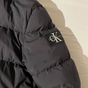 Svart dunjacka från Calvin Klein - Säljer en snygg svart dunjacka från Calvin Klein i storlek M. Jackan har en pufferstil med dragkedja och huva, perfekt för kalla vinterdagar. Den är i mycket bra skick och har en stilren design med CK-logga på ärmen. Supervarm och bekväm!  Priset kan diskuteras 