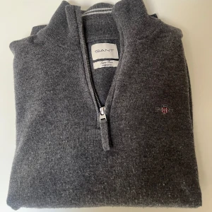 Grå ulltröja från GANT - Säljer en stilren grå tröja från GANT i superfin lammull. Den har en praktisk dragkedja framtill och en liten broderad logga på bröstet. Perfekt för kyliga dagar och passar både till vardags och lite finare tillfällen. 🐑✨Lite nopprig men går att lösa med en kam. Pris kan diskuteras vid snabbaffär!!