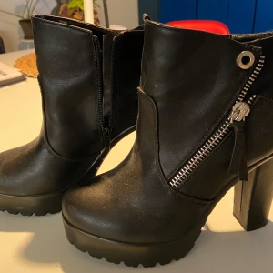 Svarta boots med dragkedja stl 38 - Snygga svarta boots med hög klack och cool dragkedja på sidan. Perfekta för höst och vinter, ger en edgy look till vilken outfit som helst. Sulan har ett stjärnmönster för extra grepp. Använd dem till fest eller en utekväll för att verkligen sticka ut!