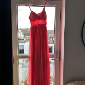 Korallröd långklänning med detaljer - Säljer en fantastisk korallrödrosa långklänning med smala axelband och v-ringning. Klänningen har fina dekorativa detaljer längs banden och en elegant passform som passar perfekt för festliga tillfällen. Materialet är mjukt och flödande, vilket ger en vacker siluett.
