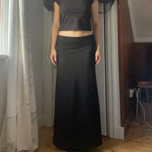 Svart långkjol  - Säljer en elegant svart långkjol i mycket bra skick. Kjolen har en tight passform upptill och faller snyggt ner mot golvet. Perfekt för både vardag och festliga tillfällen. Den är gjord i ett mjukt och bekvämt material som ger en smickrande siluett. Passar perfekt till en croppad topp eller en stickad tröja.