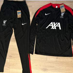 Säljer ett liverpool träningsställ med röda detaljer på axlarna. Tröjan har L.F.C.-logga och AXA-tryck på framsidan. Perfekt för träning eller vardagsbruk. Helt ny med taggar kvar.