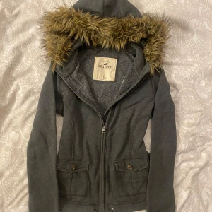 Hollister kofta med päls  - Säljer en snygg grå kofta från Hollister med en mysig pälsluva. Kofta  har dragkedja och två fickor med knappar framtill. Perfekt för höst och vinter när du vill hålla dig varm. Storleken är dam L men passar som M. Den är vintage dvs från 2000-talet!💕