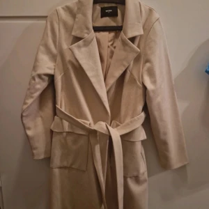 Beige kappa från Bik Bok - Säljer en stilren beige kappa från Bik Bok i storlek M. Den har en klassisk design med knytbälte i midjan och långa ärmar. Perfekt för höst och vår, och ger en elegant touch till vilken outfit som helst. Kappan är i mycket bra skick och redo för nya äventyr!