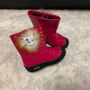 Rosa vinterstövlar med kattdetalj - Supercoola rosa vinterstövlar med en söt kattdetalj och fluffig päls runt skaftet. De har en dragkedja på sidan för enkel på- och avtagning. Perfekta för att hålla fötterna varma och stiliga under kalla dagar. Sulan är robust och ger bra grepp på isiga underlag.  Stlk kvar  27.29,30,31.                                        Med fleece inuti, rekommenderar att välja en Stlk större 