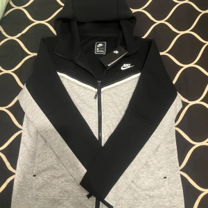 Nike tech - Säljer en snygg grå och svart hoodie från Nike i storlek M. Den har en dragkedja framtill och en justerbar huva. Perfekt för både träning och vardag. Helt ny med taggar kvar!