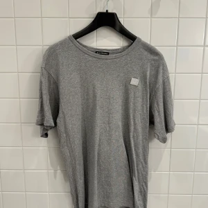 Grå t-shirt från Acne Studios i storlek L - Säljer en stilren grå t-shirt från Acne Studios i storlek L. Den är gjord av mjuk bomull och har en liten broderad detalj på bröstet. Perfekt för en avslappnad look, oavsett om det är till vardags eller en chill fest. T-shirten är i mycket bra skick och redo för en ny ägare!