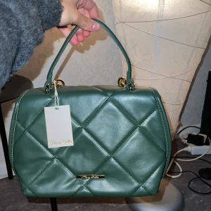 Oanvänd grön handväska från Flora Bella - Snygg grön handväska i quiltat mönster från Flora Bella. Väskan har en elegant guldkedja och en praktisk dragkedja inuti med axelrem. Perfekt för både vardag och fest! 👜✨