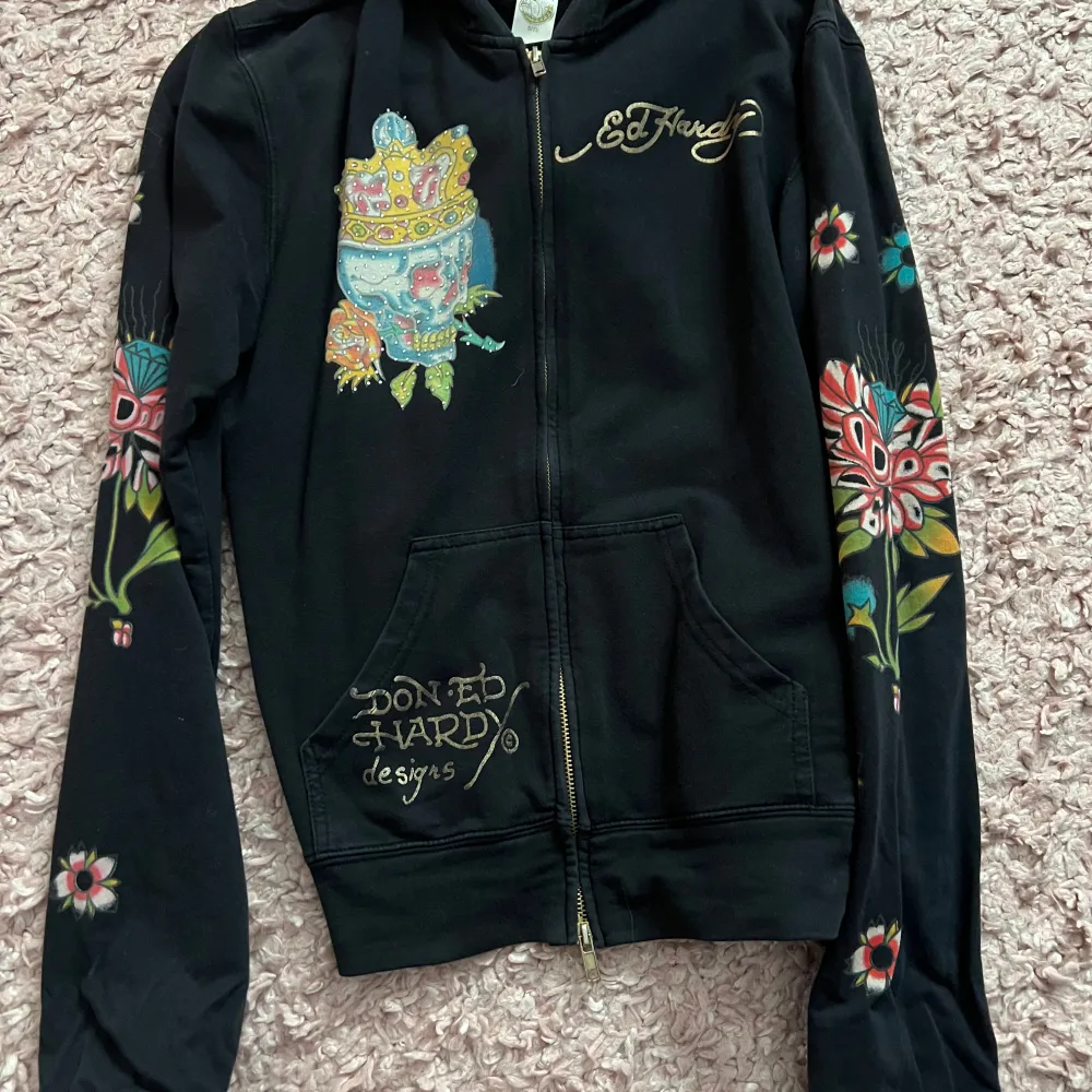 Säljer en cool svart hoodie från Ed Hardy med färgglada broderade motiv av en dödskalle med krona och blommor. Den har dragkedja fram och långa ärmar med mönster. Perfekt för en unik och stilfull look! Skulle själv säga att den är lite 2000s vibe. Xl I barnstorlek som motsvarar s I vuxen storlek. Det är bara att skriva om du har några frågor 💕💕. Hoodies.