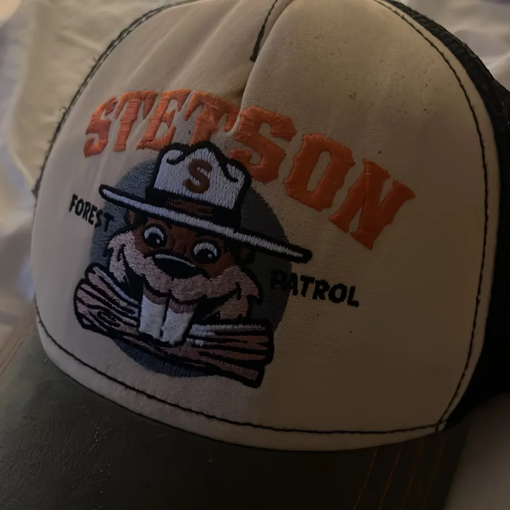 Säljer en cool Stetson keps med ett skogspatrullmotiv. Kepsen har en beige framsida med ett tryck av en bäver och texten 'Forest Patrol'. Baksidan är i svart mesh för bra ventilation. Perfekt för sommarens äventyr!. Accessoarer.