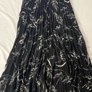 Svart och vit blommig kjol - Säljer en superfin svart kjol med vita blommönster. Kjolen är plisserad och har en härlig flowy känsla, perfekt för både vardag och fest. Den är i mycket bra skick och passar perfekt till både sommar och vår. Kjolen är i en längre modell och har en elastisk midja för extra komfort.