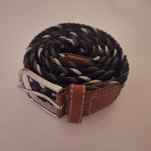 Flätat bälte - Säljer ett snyggt flätat bälte i blått, grönt och vitt med bruna läderdetaljer. Bältet har en silverfärgad spänne och är perfekt för att ge en stilren touch till din outfit. Passar både till vardags och fest. Perfekt för alla säsonger!