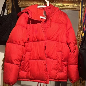 Röd dunjacka med huva hm  - Säljer en superfin röd dunjacka med huva. Den är perfekt för kalla vinterdagar och har en mysig, puffig stil. Jackan har långa ärmar och stängs med dragkedja. Den är i bra skick och redo för nya äventyr!