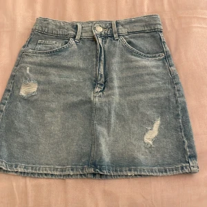 Jeanskjol i blått - Säljer en snygg jeanskjol i blått med slitna detaljer. Kjolen har en klassisk femficksdesign och stängs med dragkedja och knapp framtill. Perfekt för en avslappnad vardagslook eller en somrig outfit. Passar bra till sneakers eller sandaler!