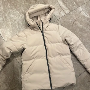 Beige dunjacka från Cross Sportswear - Säljer en snygg beige dunjacka från Cross Sportswear i storlek XS. Jackan har en dragkedja framtill och är perfekt för kalla dagar. Den är långärmad och har en mysig huva för extra värme. Perfekt för vintern! ❄️