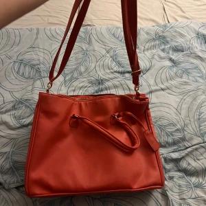 Röd axelväska med dragkedja - Säljer en snygg röd axelväska i syntetmaterial. Väskan har en praktisk dragkedja och flera fack inuti för enkel organisering. Perfekt för både vardag och fest! 👜