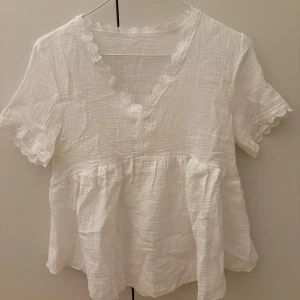 Vit blus med spetsdetaljer - Säljer en söt vit blus med v-ringning och spetsdetaljer längs halsen och ärmsluten. Blusen har en luftig passform och är perfekt för vår och sommar. 