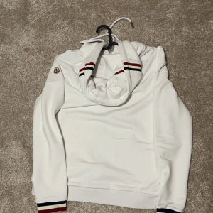 Vit hoodie från Moncler - Säljer en snygg vit hoodie från Moncler i mycket bra skick. Den har en dragkedja framtill och en justerbar huva med röd och blå randig detalj. Perfekt för både höst och vår. Moncler-loggan sitter på ärmen. Passar perfekt för en stilig look. 