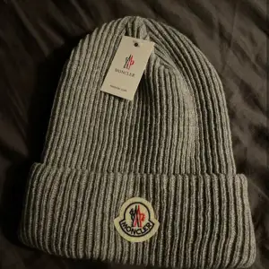 Säljer en snygg grå ribbad mössa från Moncler. Den är perfekt för kalla dagar och har en klassisk design med Moncler-loggan framtill. Mössan är i nytt skick och passar perfekt till både vardag och vinteraktiviteter.